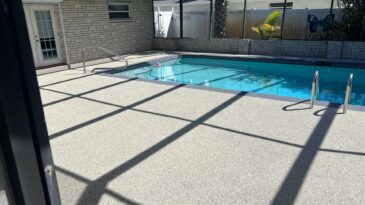 Screened-in backyard pool with steps and handrails, surrounded by textured deck. A brick wall and glass doors are in the background, with palm trees and a butterfly decoration near the fence.