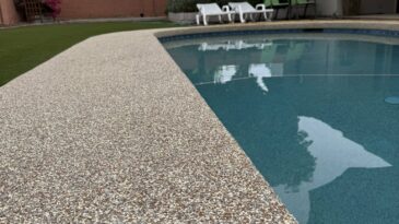 Close-up of a speckled pool deck beside a clear blue swimming pool in a backyard, with lounge chairs, green lawn, trees, and a brick wall visible in the background.