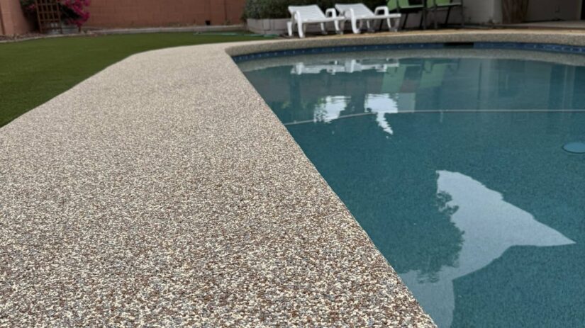 657629519_122152811762979862_4016699787727911207_n Close-up of a speckled pool deck beside a clear blue swimming pool in a backyard, with lounge chairs, green lawn, trees, and a brick wall visible in the background.