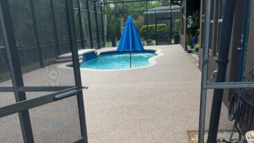 A screened-in patio with a pool, tan-speckled flooring, and a closed blue umbrella at the pools center. Greenery and patio furniture are visible in the background.