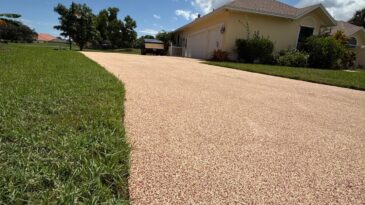 A wide, tan-colored driveway leads to a light yellow house with a garage, bordered by green grass and trees under a bright blue sky with fluffy white clouds.