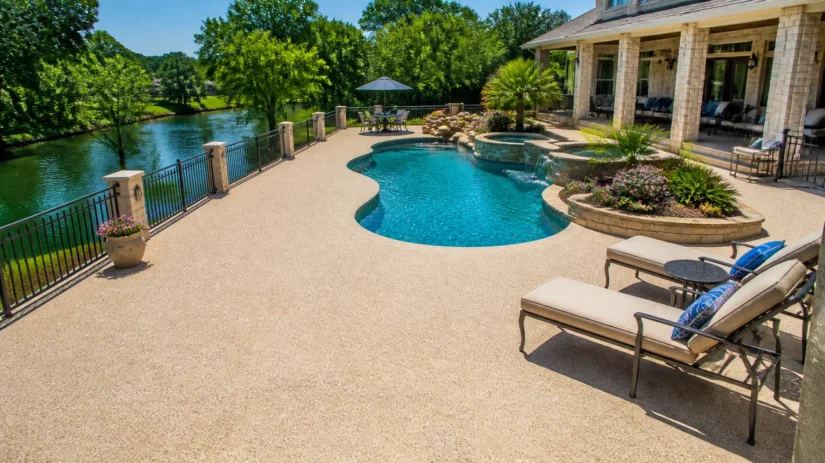 J2GLNvck9N A luxury backyard pool with lounge chairs, a hot tub, landscaping, and a view of a river bordered by trees, next to a large stone house with a shaded patio.