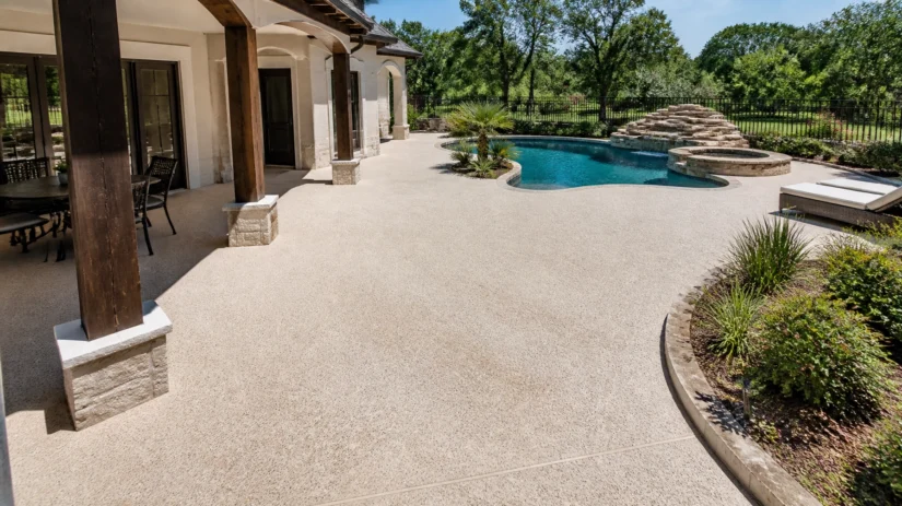 Lfe60-KsTZU Spacious backyard patio with beige stone flooring, covered seating area, and a curvy pool featuring a stone waterfall. Green trees and shrubs surround the pool area, with lounge chairs to the right.