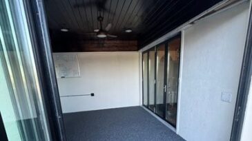 A room with glass doors, dark wood ceiling, ceiling fan, and gray speckled carpet, viewed from an open doorway. The walls are white and there is a map or picture on one wall.